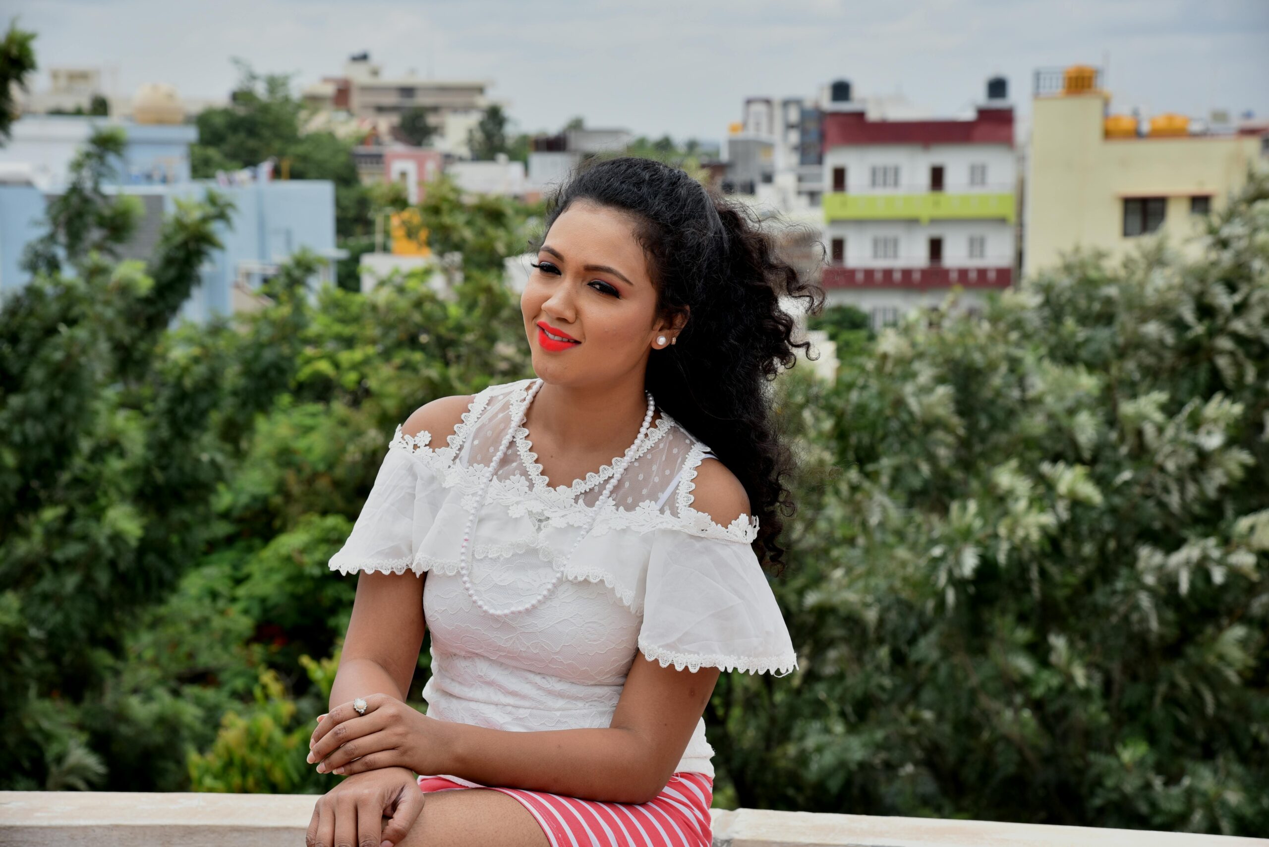 A cheerful woman sitting outdoors in a white lace top against a cityscape backdrop.